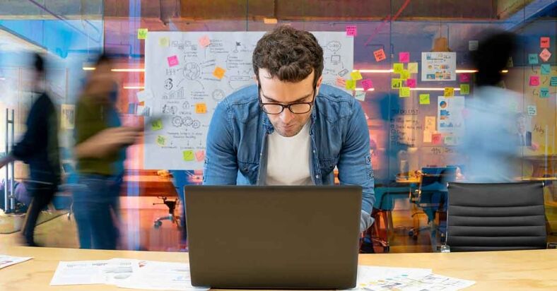 Man working at a creative office using his computer and people moving in the background Man working at a creative office using his computer and people moving in the background