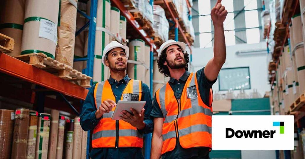 Young caucasian worker showing shelves to his african manager in a warehouse holding digital tablet with hardhat and safety vest Two workers in a warehouse holding digital tablet with hardhat and safety vest
