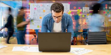 Man working at a creative office using his computer and people moving in the background