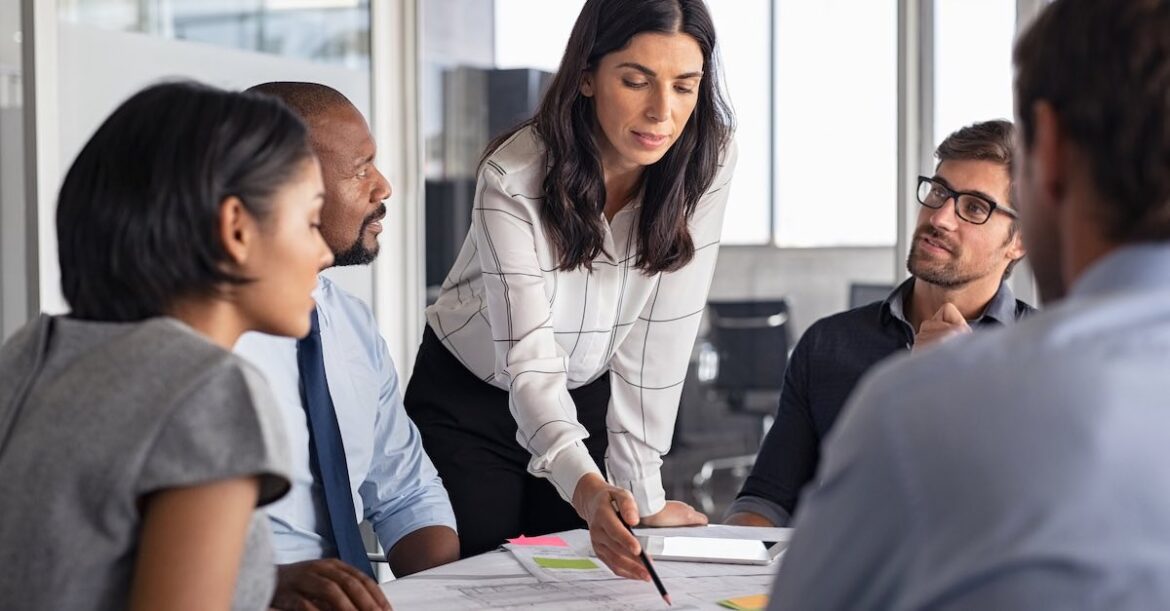 Female project manager leaning over a table of papers with her team paying attention.